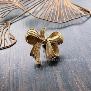 May include: A gold-toned metal bow tie shaped like a bow. The bow is textured and has a slightly worn look.