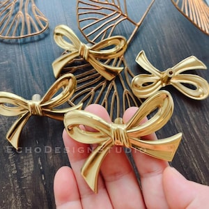 May include: Four gold-toned bow-shaped cabinet knobs. The knobs are made of metal and have a shiny finish. The bows are tied in a knot at the center.