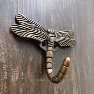 Könnte beinhalten: Ein goldfarbener Libellenhaken mit detailliertem Design. Der Haken ist an einer dunklen Holzoberfläche befestigt.