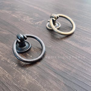Peut inclure: Deux poignées décoratives en forme d'anneau, l'une noire et l'autre dorée, avec un anneau rond et un bouton décoratif. Les poignées sont sur une surface en bois.