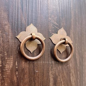 May include: A pair of antique bronze door knockers. Each knocker features a floral backplate and a circular handle. They are mounted on a dark wooden surface. The text "ECHODESIGNSTUDIO" is visible.