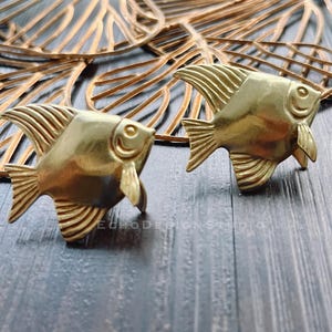 May include: Two gold-coloured metal fish figurines with detailed scales and fins. The fish are facing different directions and are on a wooden surface.