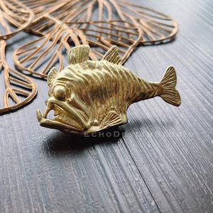 Könnte beinhalten: Ein goldfarbener Metallfisch-Anhänger mit einer detaillierten, strukturierten Oberfläche. Der Fisch hat einen großen, offenen Mund und einen langen, schlanken Körper.