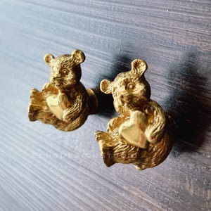 Peut inclure: Deux poignées de tiroir en forme d'ours dorées avec un motif en forme de cœur. Les ours tiennent les cœurs dans leurs pattes.