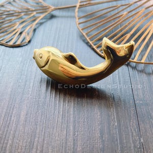 May include: A gold-coloured fish-shaped door handle or pull. The handle is on a dark wood surface.