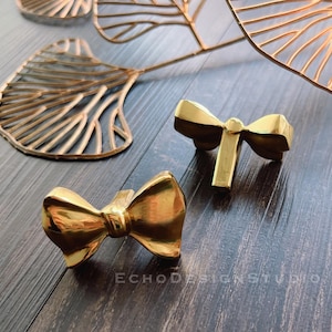 May include: Two gold-colored bow-shaped cabinet knobs. The knobs are made of a shiny metal and are positioned on a dark wood surface. The image also shows decorative gold leaf-shaped objects in the background. The text "ECHO DESIGN STUDIO" is visible.