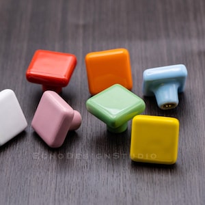 Op de afbeelding: Assortiment van vierkante keramische kastknoppen in verschillende kleuren, waaronder wit, roze, oranje, rood, lichtblauw, groen en geel.