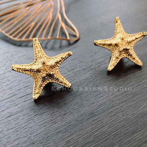 May include: A pair of gold-tone starfish earrings. The earrings are made of metal and have a textured surface.