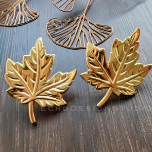 Op de afbeelding: Twee gouden metalen esdoornbladeren. De bladeren zijn gedetailleerd en hebben een licht getextureerd oppervlak.