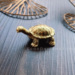 Könnte beinhalten: Eine kleine goldfarbene Schildkrötenfigur. Die Schildkröte ist aus Metall und hat ein detailliertes Design.