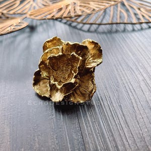 Puede incluir: Una flor de metal de color dorado con una superficie texturizada. La flor está compuesta por varios pétalos y tiene un aspecto realista.