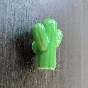 Può includere: Una statuetta di cactus in ceramica verde chiaro lucido. Il cactus ha un design tridimensionale con più braccia e una superficie liscia e riflettente. La base è piatta e la statuetta è posta su uno sfondo scuro con venature del legno. Il testo "ECHODESIGNSTUDIO" è visibile.