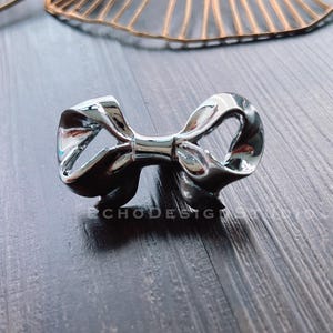 Könnte beinhalten: Ein silberner, bogenförmiger Schrank- oder Schubladenknopf. Der Bogen ist aus Metall und hat eine glatte, polierte Oberfläche.