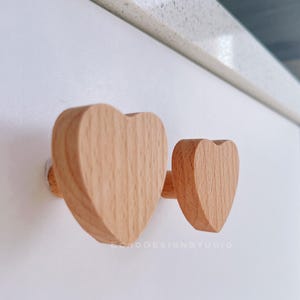 May include: Two wooden heart-shaped hooks mounted on a white wall. The hooks are made of light brown wood and have a natural finish.