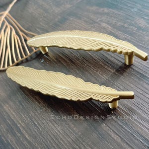 May include: Two gold-toned metal feather-shaped cabinet pulls. The pulls have a detailed feather design and are approximately 10 cm long.