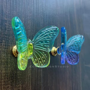 May include: Two butterfly-shaped cabinet knobs. The knobs are made of transparent glass with a gradient of colours, including blue, green, and yellow. The base of each knob is gold-coloured. The knobs are mounted on a dark wood surface.