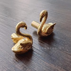 Peut inclure: Deux boutons de meuble dorés en forme de cygne. Les boutons présentent des détails de plumes texturées et des cous incurvés. Les boutons sont en métal et sont posés sur une surface en bois foncé.