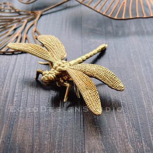 May include: A gold-coloured metal dragonfly figurine with intricate details. The dragonfly is perched on a dark wooden surface.