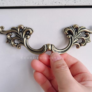May include: Ornate antique brass drawer pull with a curved handle and decorative floral detailing. The hardware is held against a white surface. The Echo Design Studio logo is visible.