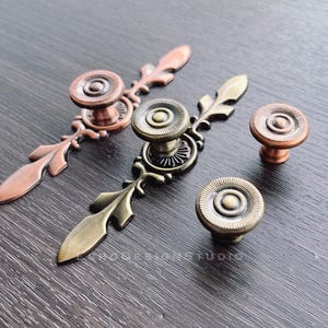 May include: Four antique-style cabinet knobs with ornate designs. Two knobs are copper with a circular design and two knobs are brass with a circular design.