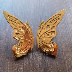May include: Two gold butterfly wing-shaped decorative objects. The wings have detailed patterns and textures, and are made of a shiny, metallic material. They are likely for home decor or artistic projects. The objects are displayed on a wooden surface.