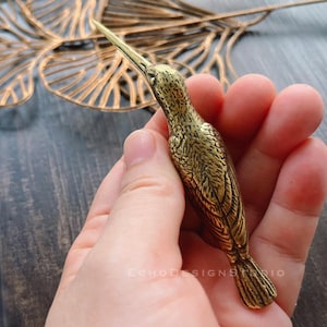 May include: A detailed, gold-coloured hummingbird figurine. The bird is crafted with intricate feather detailing and a long, pointed beak. The figurine is held in a hand, showcasing its size and craftsmanship. The Echo Design Studio logo is visible.