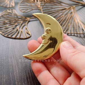 May include: A gold crescent moon shaped pin with a smiling face. The pin is made of metal and has a shiny finish.