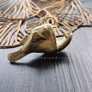 May include: A gold-colored metal sculpture of a anglerfish with its mouth open and teeth visible. The sculpture is on a dark wood surface.