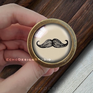 Può includere: Un pomello rotondo per armadio in ottone con un inserto in vetro che presenta un'illustrazione in bianco e nero di un baffo.