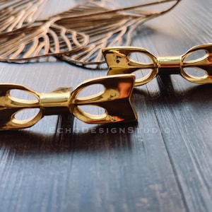 May include: Two gold-colored bow-shaped cabinet pulls. The pulls have a polished finish and a symmetrical design with open loops. The background includes a decorative gold metal piece and a dark wood surface.