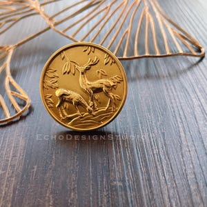 May include: Gold-toned metal coin with a raised design of two deer standing in a forest. The coin is on a wooden surface.