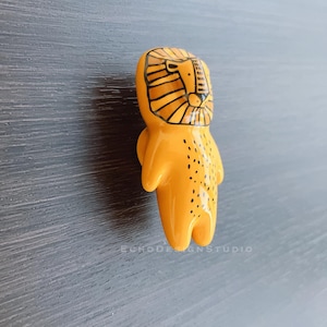 May include: A yellow ceramic lion figurine with black details. The lion has a stylized face with a mane and spots on its body.