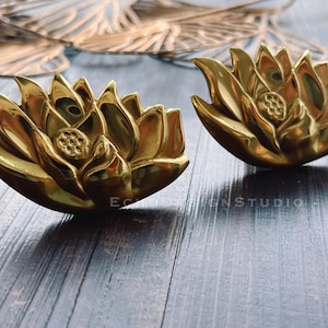 以下が含まれることがあります： 金色の蓮の花の彫刻が2つ。花びらのデザインが細かく表現されています。彫刻は光沢のある金属製で、暗い木製の表面に置かれています。全体的なテーマは装飾芸術です。