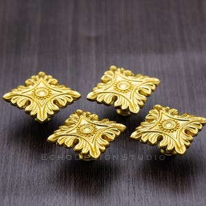 Peut inclure: Quatre boutons décoratifs pour armoire de couleur or avec une forme carrée et des motifs floraux complexes. Chaque bouton présente un cercle central avec un motif en relief.