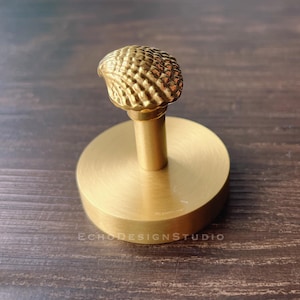 Puede incluir: Un gancho de metal dorado con un diseño texturizado en forma de concha. El gancho está montado en una base redonda.