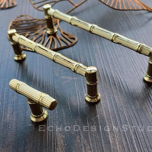 Può includere: Tre maniglie per armadi dorate con un design a bambù. Le maniglie sono di dimensioni diverse, la più grande misura circa 15 cm di lunghezza.