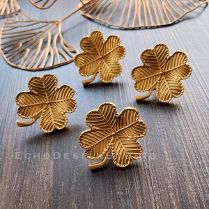 May include: Four gold-colored four-leaf clover shaped metal ornaments on a wooden surface.