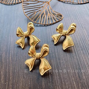 May include: Three gold-colored bow-shaped decorative pieces. The bows are made of metal and have a shiny finish.