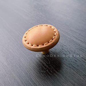 May include: A round, tan leather cabinet knob with a stitched edge and a brass base.