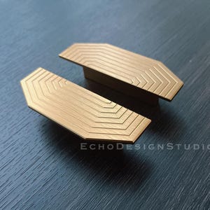 May include: Two gold-toned metal cabinet pulls with a geometric pattern. The pulls are shaped like a rounded rectangle with a slightly curved edge.