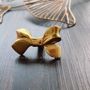 Pode incluir: Uma maçaneta de armário ou puxador de gaveta em forma de laço de cor dourada. O laço é feito de metal e tem um acabamento brilhante.