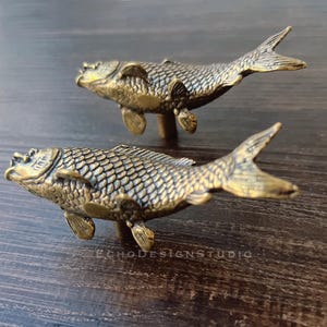 Peut inclure: Deux poignées de meubles en forme de poisson de couleur bronze. Les poissons ont des écailles, des nageoires et des queues détaillées. Elles sont montées sur de petits poteaux pour être fixées aux meubles. Les poignées sont posées sur une surface en bois foncé.