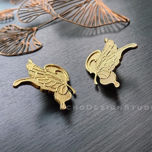 May include: Two gold butterfly-shaped drawer pulls with intricate details. The pulls are on a dark wood surface.
