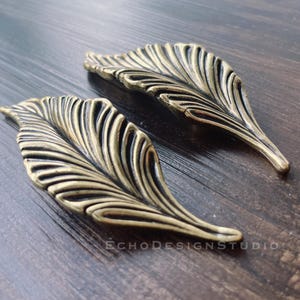 Puede incluir: Dos tiradores de cajón en forma de hoja de color latón antiguo. Los tiradores tienen vetas detalladas y un diseño curvo. El fondo es una superficie de madera oscura. Los tiradores son de EchoDesignStudio.