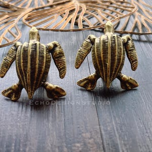 May include: Two bronze-colored turtle figurines with detailed shell patterns are displayed on a dark wooden surface. The turtles are positioned facing forward, with their flippers extended. A decorative gold leaf-like object is in the background. The text "ECHODESIGNSTUDIO" is visible.