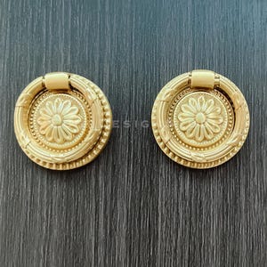 以下が含まれることがあります： 花柄の金色のキャビネットノブが2つ。ノブにはリングプルが付いており、装飾が施されています。