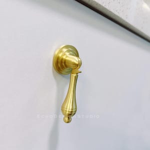 Op de afbeelding: Een gouden kastknop met een traanvormig handvat. De knop is gemonteerd op een wit oppervlak.