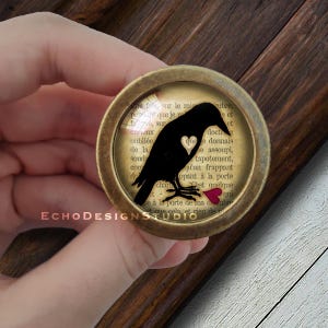 May include: A black crow silhouette holding a red heart in its beak, framed in a gold-toned round pendant with a vintage book page background. The text on the page reads "...que je m'efsur le miubre, efsur..."