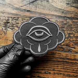 K&ouml;nnte beinhalten: Schwarzer, gestickter Aufn&auml;her mit einem Augendesign in Wolkenform. Das Auge und die Wolkenkontur sind wei&szlig; auf schwarzem Hintergrund gestickt. Der Aufn&auml;her hat eine tropfenf&ouml;rmige Form unter dem Auge.