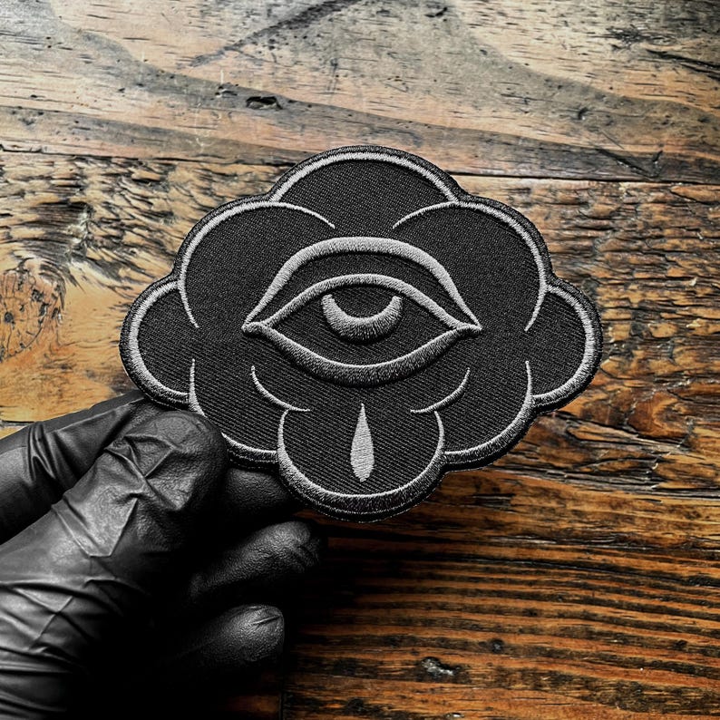 K&ouml;nnte beinhalten: Schwarzer, gestickter Aufn&auml;her mit einem Augendesign in Wolkenform. Das Auge und die Wolkenkontur sind wei&szlig; auf schwarzem Hintergrund gestickt. Der Aufn&auml;her hat eine tropfenf&ouml;rmige Form unter dem Auge.